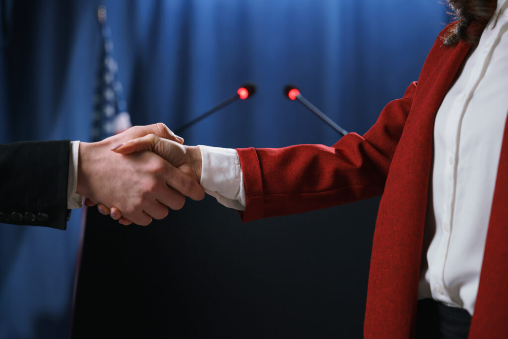 White American politicians shaking hands at the debates, looking in camera, standing on a blue background with American flag