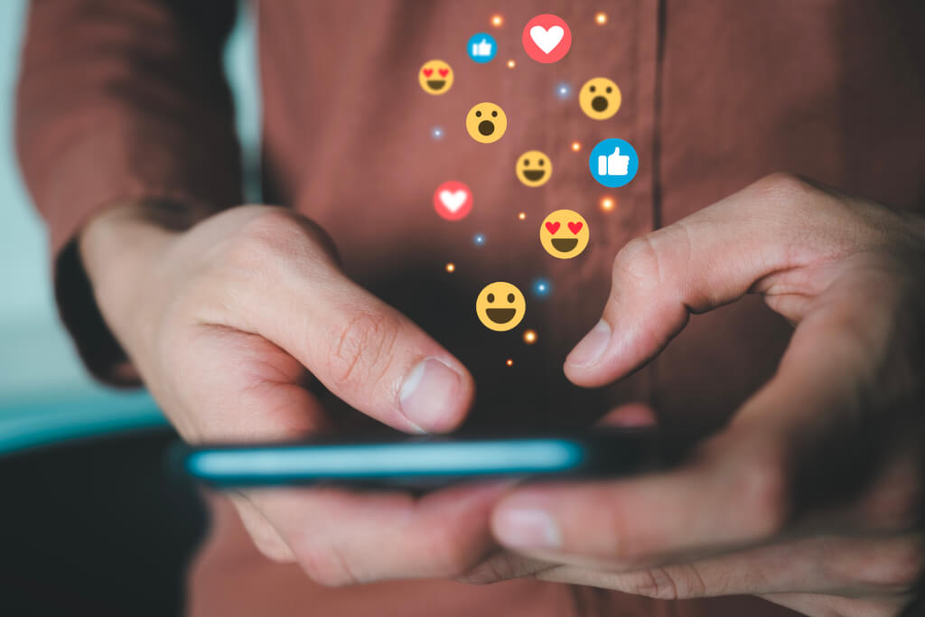 Close up of male hands holding smartphone connecting with people online using social media with illustration hologram of emojis showing and floating.