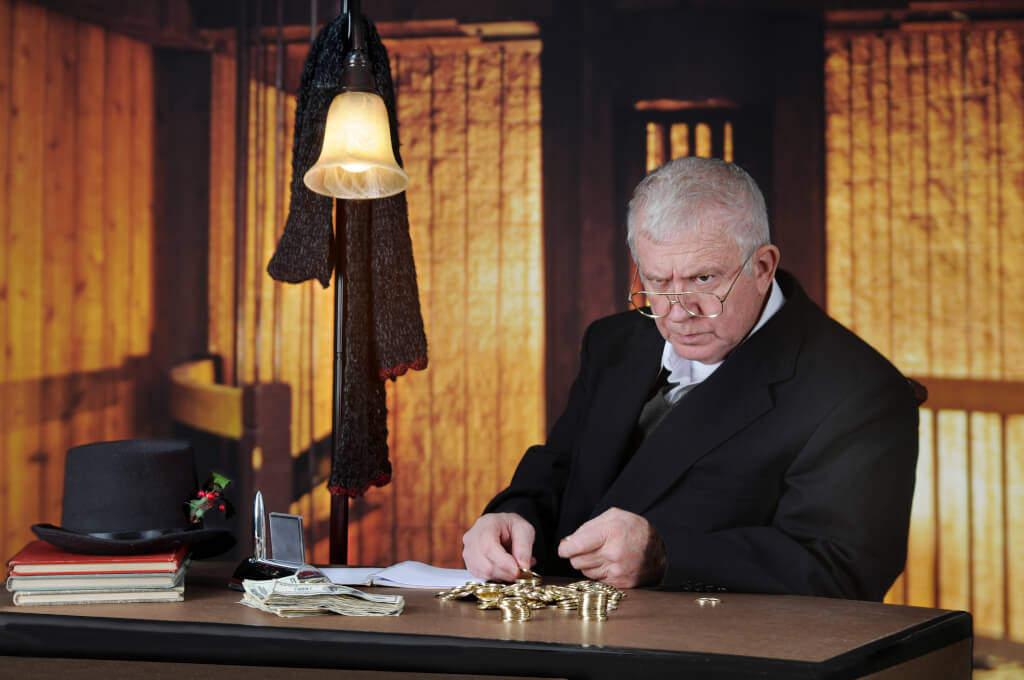 A grumpy old miser sitting at his desk counting gold coins by a stack of big bills.