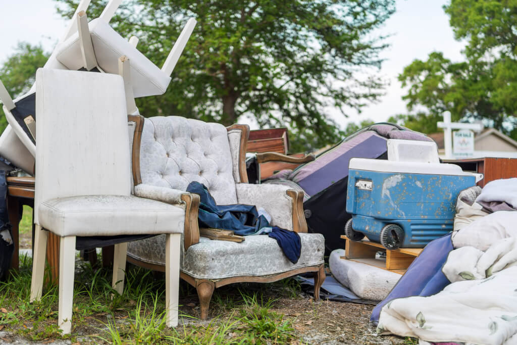 A cluttered collection of household items, including upholstered chairs and a vintage cooler, sits abandoned on a suburban curb, illustrating themes of waste, moving day, or garage clearance.